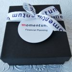 Square black matte box with a white printed ribbon and a circular branding tag.