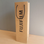 Tall, narrow-format natural kraft paper bag featuring a bold vertical black silkscreen logo and black cord handles.