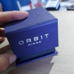 Small blue textured hinged lid box with silver foiled branding for rings.
