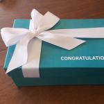 Teal-colored rigid gift box decorated with a wide white satin ribbon and bow.