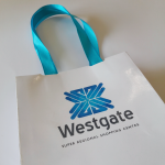 White high-gloss laminated paper bag featuring wide turquoise satin ribbon handles and multi-tonal blue branding
