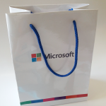 Small white high-gloss paper bag with blue braided rope handles and multi-colored brand striping