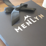 Black matte-finish paper bag with a silver foil logo and a premium black satin ribbon handle tied in a bow.