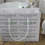 Landscape-format white paper bag with repeating logo pattern and white cord handles