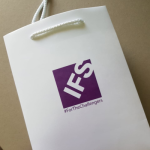 White gloss-laminated paper bag featuring a purple square logo and white braided cord handles