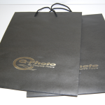 Pair of large black matte-finish paper bags featuring silver foil-stamped logos and black cord handles.
