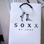Small white matte-finish paper bag featuring a black silkscreened logo and black braided handles.