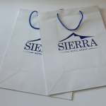Two white matte-finish paper bags featuring a large blue silkscreened logo and blue cord handles.
