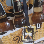 Custom-printed cardboard carry box holding six glass bottles with an attached marketing tag.