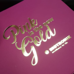 High-shine gold foil logo on a matte black rigid substrate or vibrant pink substrate.