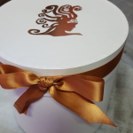 Large white circular hat box with a copper foil logo and matching satin ribbon.