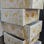 Cream rigid box flats undergoing a gold silk-screening process for branding and finished white boxes with metallic gold illustrations.